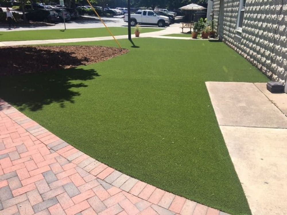 Artificial turf landscaping with a paved path and outdoor seating area in a sunny park.