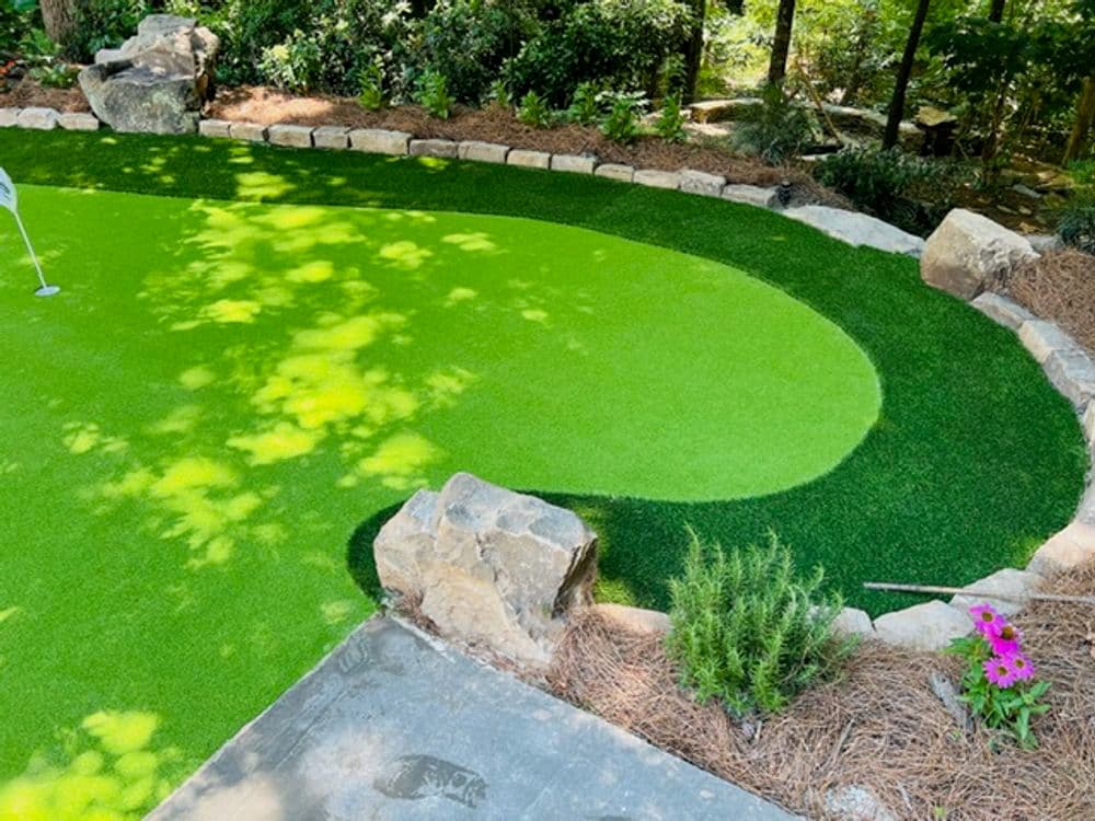 Artificial turf putting green with stone border and landscaped edges in a garden setting.