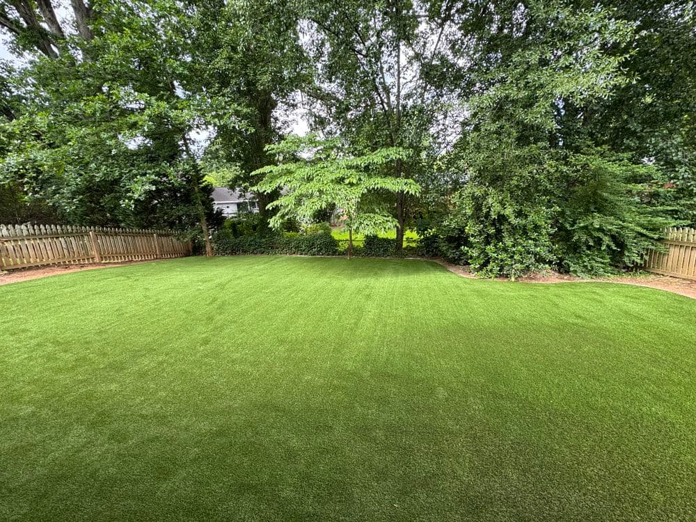Lush green artificial grass lawn surrounded by trees and a wooden fence.