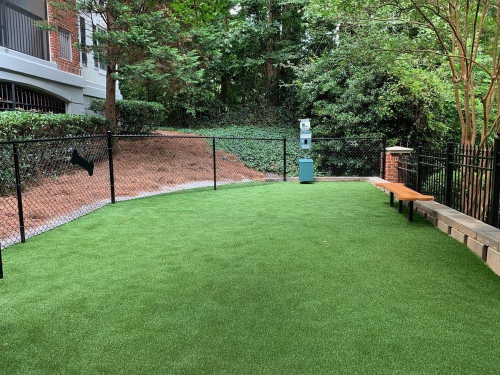 Dog park with artificial turf, fenced area, bench, and greenery surrounding it.