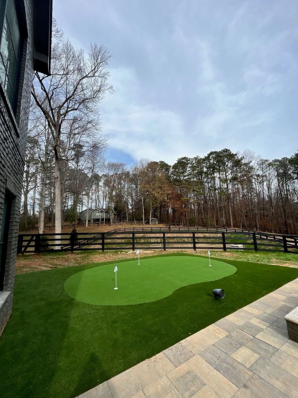 Home backyard putting green with synthetic grass and black wooden fence in a wooded area.