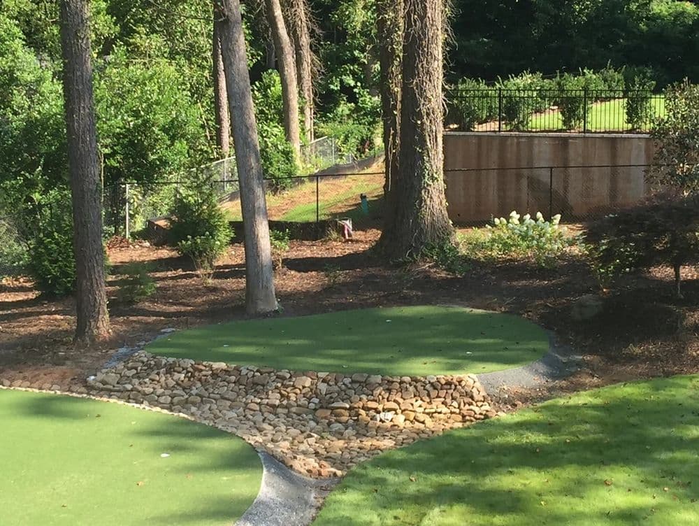 Golf putting green surrounded by trees and landscaping features in a backyard setting.