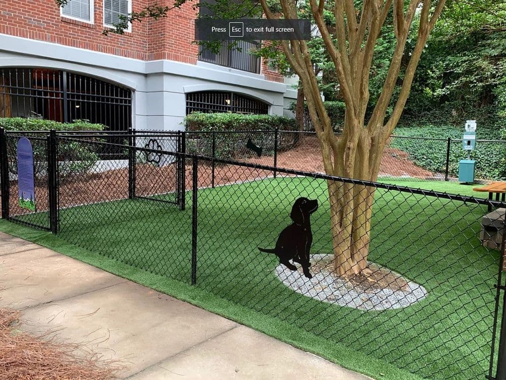 Dog park with a fenced area, tree, and grass, featuring a wall-mounted dog silhouette.