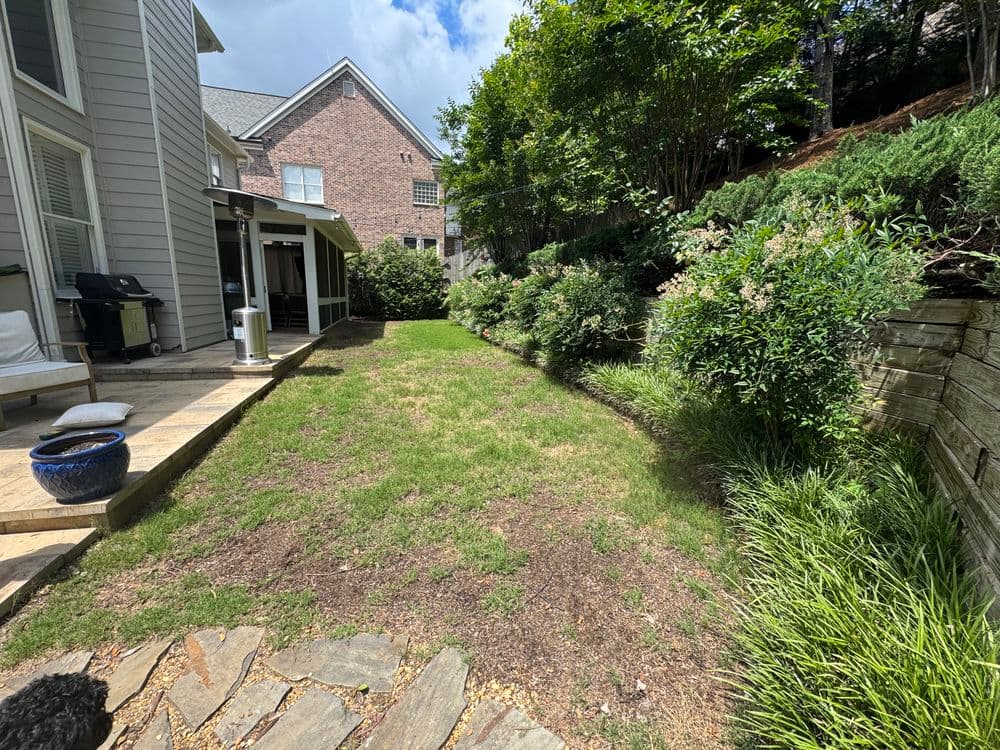 Backyard landscape featuring green grass, shrubbery, and stone path next to a home.