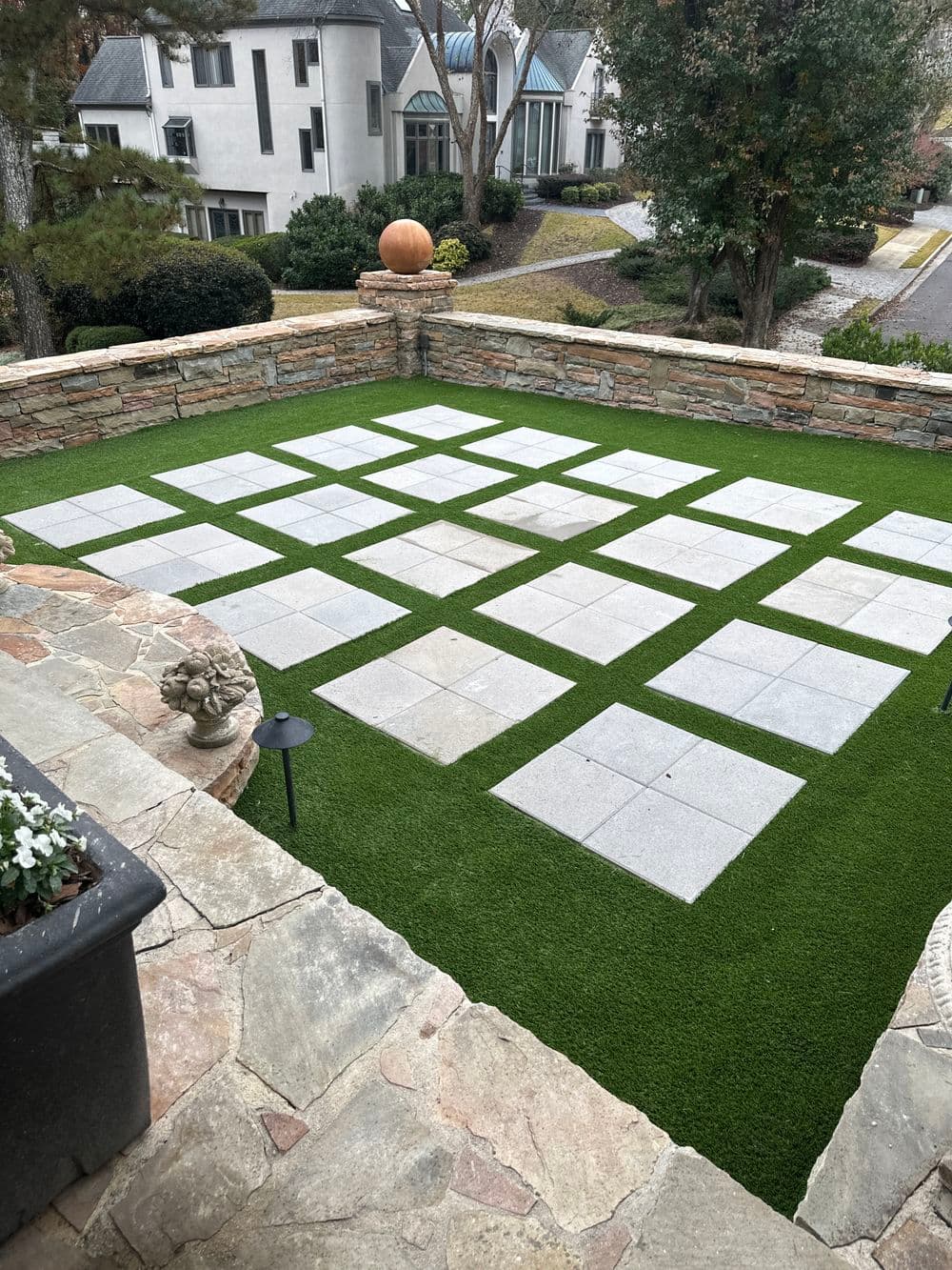 Modern patio design with stone pavers on artificial grass, surrounded by landscaped greenery.