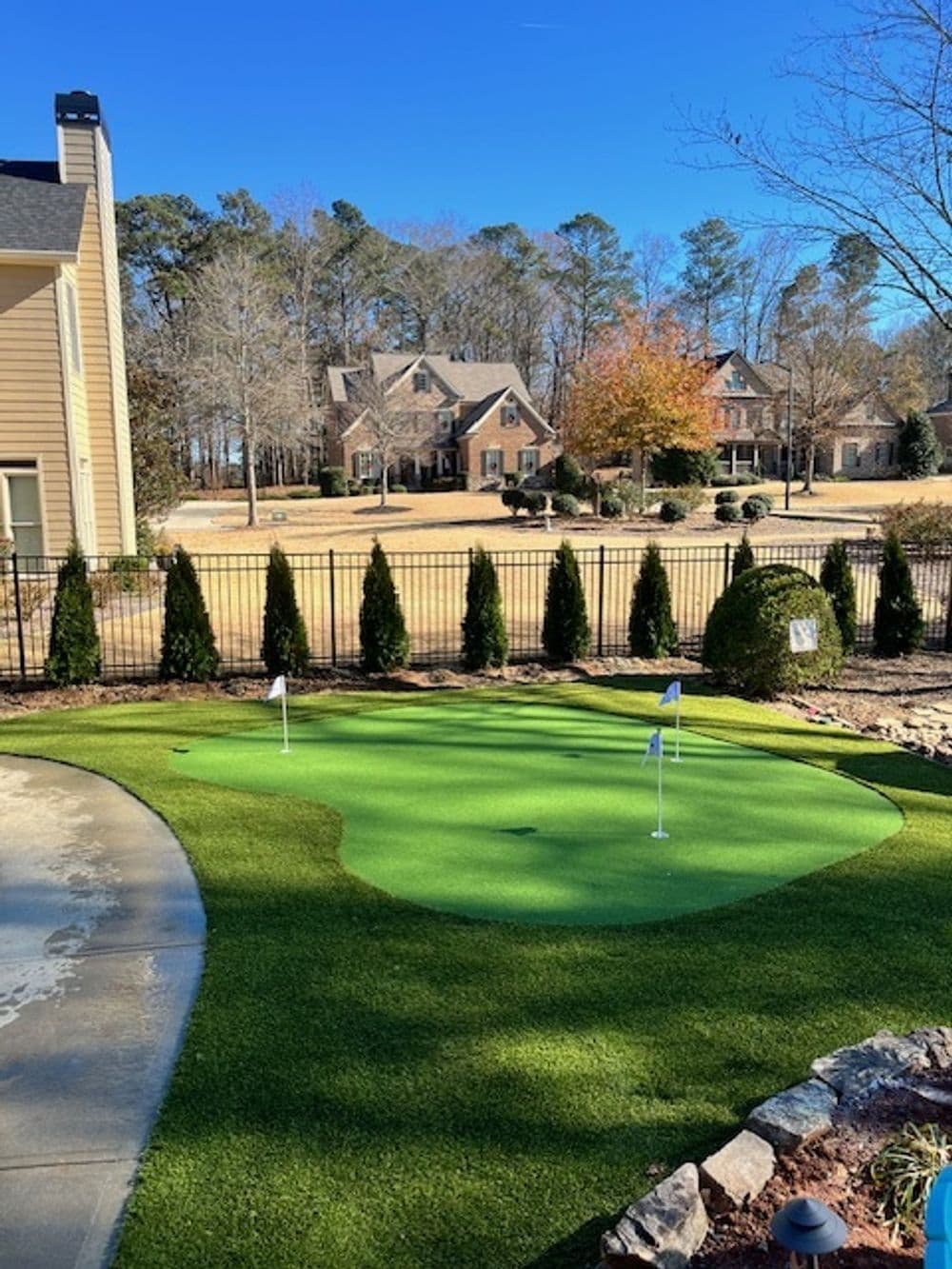 Backyard putting green with flags, framed by trees and decorative shrubs under clear blue sky.