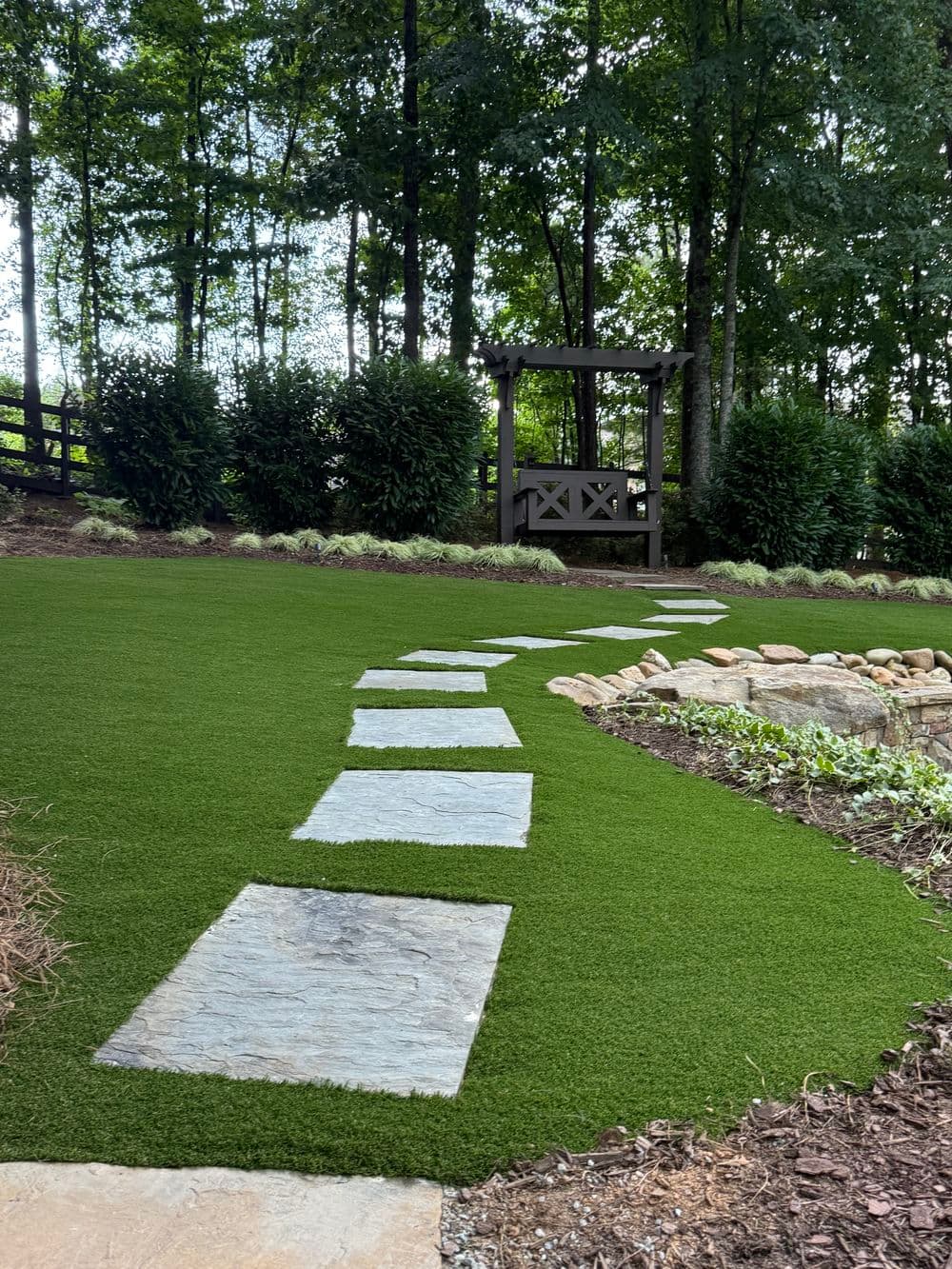 Curved stone path on artificial grass leads to a wooden gazebo in a lush garden setting.