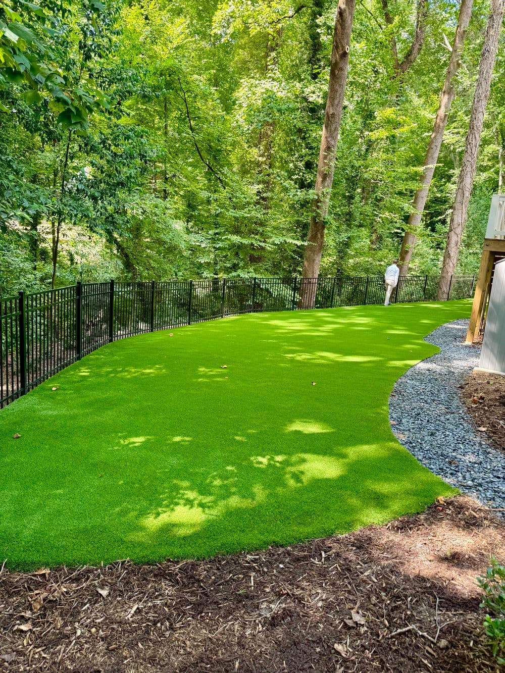 Lush green artificial turf installation in a wooded backyard with a black fence.