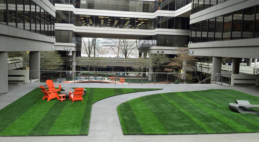 Modern office courtyard with artificial grass, orange chairs, and architectural buildings.